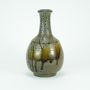 May include: A ceramic vase with a textured surface and a dark brown and olive green glaze. The vase has a narrow neck and a bulbous body with a drip effect. The vase is likely used for decorative purposes.