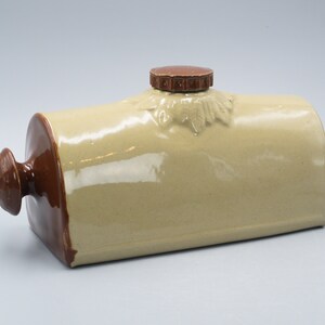 Può includere: Una bottiglia di acqua calda in ceramica con un coperchio e un manico marroni. La bottiglia è beige e ha una finitura liscia e lucida.