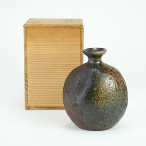 Puede incluir: Un jarrón de cerámica redondo de color marrón oscuro con un cuello estrecho y una superficie texturizada. El jarrón tiene un aspecto moteado con tonos verdes y marrones. Detrás del jarrón hay una caja de madera.