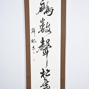 May include: A vertical scroll with black calligraphy on white paper. The text is in a traditional East Asian style, with vertical columns of characters. The scroll has a brown border and a decorative top edge.