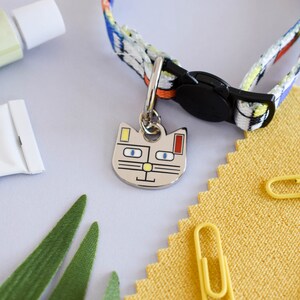 Puede incluir: Una etiqueta de identificaci&oacute;n para mascotas con forma de gato de colores con ojos azules, nariz amarilla y detalles rojos y amarillos cuelga de un collar de gato con estampado colorido con una hebilla negra.