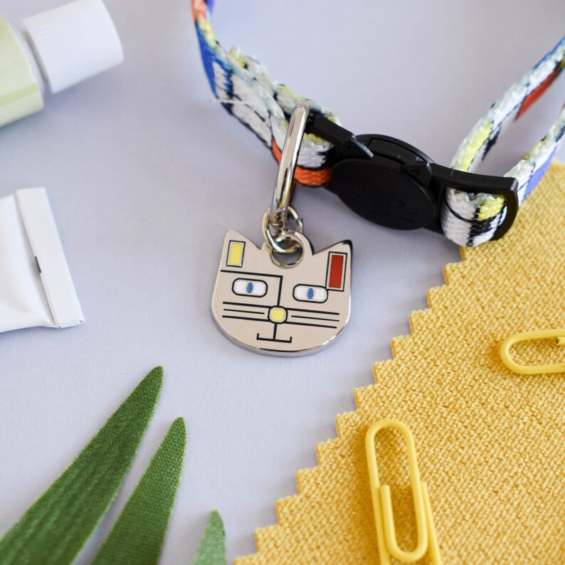 Puede incluir: Una etiqueta de identificaci&oacute;n para mascotas con forma de gato de colores con ojos azules, nariz amarilla y detalles rojos y amarillos cuelga de un collar de gato con estampado colorido con una hebilla negra.