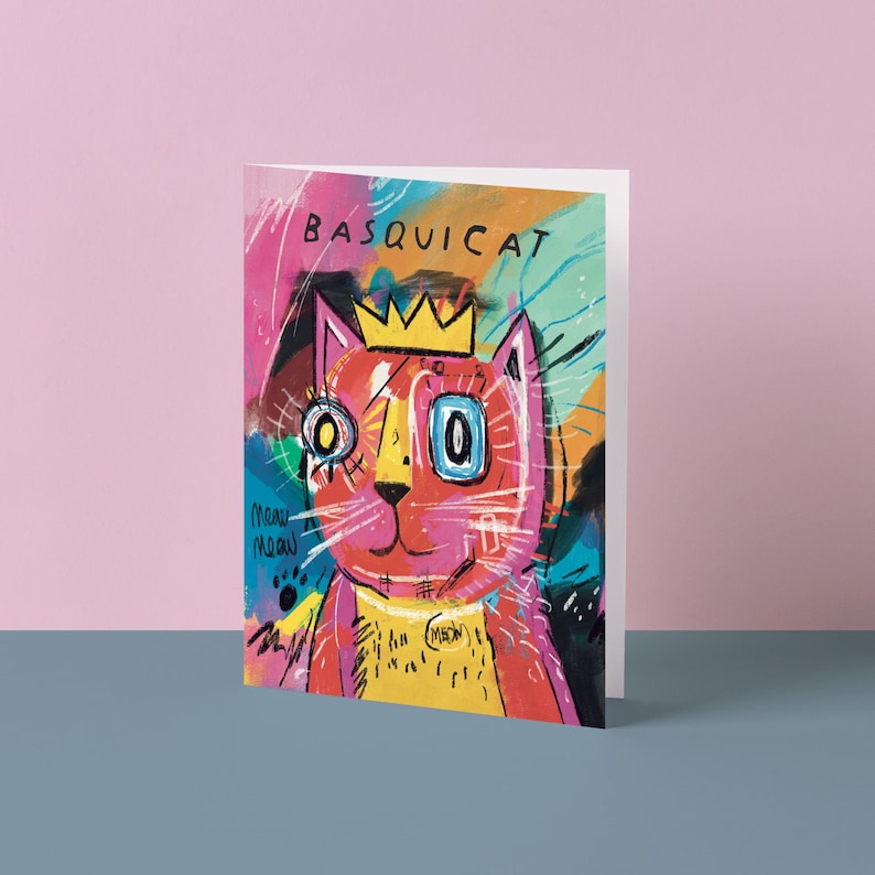 May include: Greeting card featuring a colorful, abstract illustration of a cat wearing a gold crown. The artwork includes the text "BASQUICAT" and "meow meow". The cat is primarily red and yellow, with blue and pink accents.