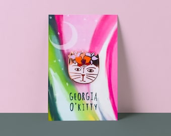 May include: A pink and green enamel pin featuring a white cat with a flower crown. The pin says "Georgia O'Kitty" on the card.