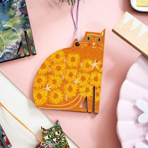 Op de afbeelding: Houten kattenornamenten in verschillende ontwerpen. Eén oranje kat heeft gele zonnebloempatronen en witte steraccenten. Andere katten hebben bloemen en abstracte ontwerpen. Elk ornament heeft een touwtje om op te hangen. De ornamenten liggen op een roze ondergrond.