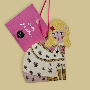 Può includere: Un ornamento in legno a forma di gatto raffigurante un personaggio dai capelli biondi con guance a forma di cuore, che indossa un indumento bianco con motivi a stella marroni. Un filo rosa lega un'etichetta quadrata rosa con la scritta Dolly Pawton e Nia Ski.