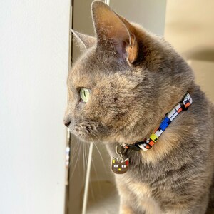 Puede incluir: Un gato atigrado gris y marr&oacute;n con ojos verdes mira por una ventana. El gato lleva un collar estampado de colores con una etiqueta en forma de gato.