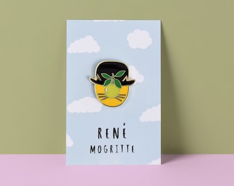 May include: A gold-edged enamel pin depicting a bowler hat with a green apple and leaves. The pin is displayed on a card with a blue sky and cloud design, and the text "RENÉ MOGRITTE" in black.