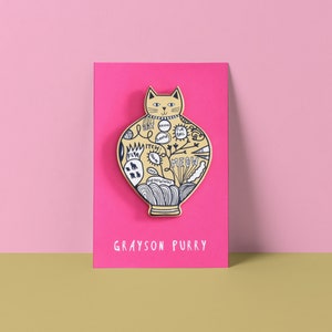 Puede incluir: Un pin amarillo con forma de gato con contornos negros y texto que dice "MEOW" y otras palabras relacionadas con los gatos. El pin está sobre un fondo rosa con el texto "GRAYSON PURRY" debajo.