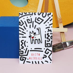 May include: A white and black pin with a cat design and a red heart, set against a white card with black abstract art. The card has the text "KEITH HAIRBALL" in red. The background includes a blue block, a yellow easel, and a paintbrush.