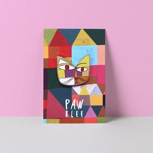 May include: A colorful enamel pin featuring a stylized cat face with geometric shapes in yellow, red, purple, orange, and white. The pin is on a card with a geometric pattern and the text "PAW KLEE".