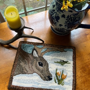 May include: A brown and white rug with a detailed image of a deer's head looking down at yellow flowers. The rug is on a wooden table with a yellow candle and a blue and white pitcher with yellow flowers.