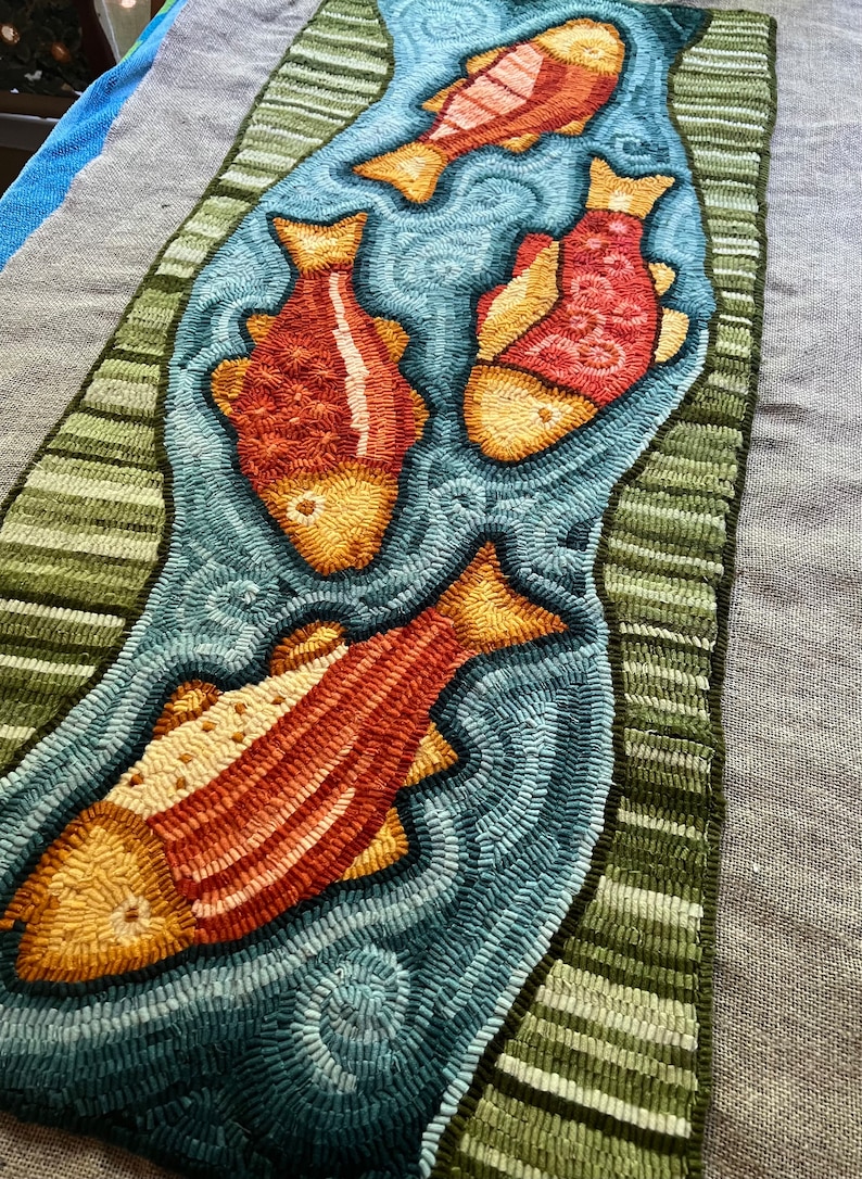 K&ouml;nnte beinhalten: Ein Teppich mit einem Design aus vier orangefarbenen und gelben Fischen, die in einem blauen Bach schwimmen. Der Bach ist von gr&uuml;nen Streifen umgeben.