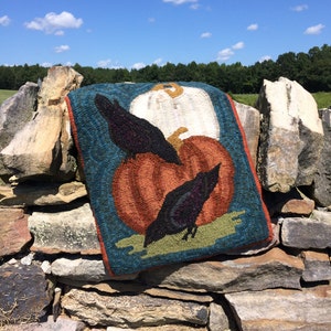 May include: A hand-stitched wool rug with a blue background features a pumpkin, two black birds, and a white gourd. The rug is displayed on a stone wall.