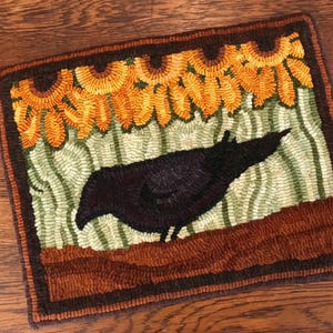 May include: Hand-hooked rectangular rug featuring a black crow silhouette against a backdrop of sunflowers and green foliage. The rug has a brown border and is made with textured yarn, creating a rustic, handcrafted appearance.
