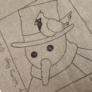 May include: A tan fabric square with a black outline drawing of a snowman wearing a hat and a bird perched on top. The snowman has a carrot nose and button eyes. The text "Woolly Woms Rugs" and "Snowman #2" are visible.