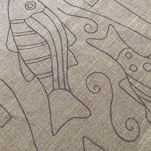 K&ouml;nnte beinhalten: Nahaufnahme eines beigefarbenen Stoffes mit einem schwarzen Stickmuster aus zwei Fischen und einem floralen Muster.