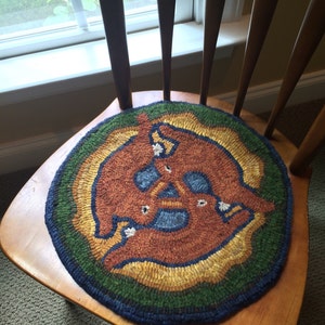 May include: A round, hand-hooked rug with a central design of three stylized brown figures. The rug features concentric rings of blue, green, yellow, and brown. It is placed on a wooden chair.