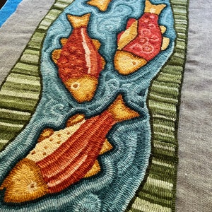 K&ouml;nnte beinhalten: Ein Teppich mit einem Design aus vier orangefarbenen und gelben Fischen, die in einem blauen Bach schwimmen. Der Bach ist von gr&uuml;nen Streifen umgeben.