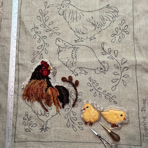May include: A textile art piece featuring a rooster and two chicks, with a ruler along the side. The rooster is rendered in brown, red, and black yarn, while the chicks are yellow. The background is a neutral linen fabric. The text "Joanne Page" is visible.