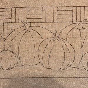May include: A brown linen fabric with a stitched design of seven pumpkins in a row. The pumpkins are outlined in brown thread and are arranged in a staggered pattern.