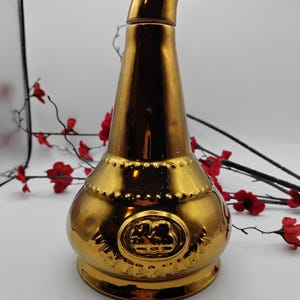 May include: A gold ceramic decanter with a unique spout design. The decanter features a raised circular emblem with a design of a ship and the text "The Famous Grouse".