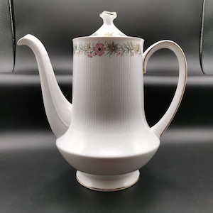 Peut inclure: Cafetière en céramique blanche avec un motif floral et des finitions dorées. La cafetière a un corps haut et fin et un bec verseur sur le côté. La poignée est incurvée et a une finition dorée.