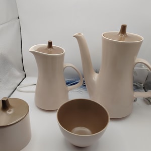 May include: A beige ceramic tea set comprising a teapot, creamer jug, sugar bowl, and a small bowl. The teapot features a long spout and a handle. The creamer and sugar bowl have lids. The set has a mid-century modern design.
