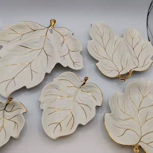 May include: Set of five white ceramic leaf-shaped serving dishes with gold trim. The dishes are arranged on a white surface.