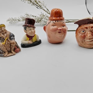 May include: Set of four ceramic figurines with whimsical faces. The figurines are painted in shades of brown, yellow, and white. One figurine is a man with a pipe, another is a man with a hat, another is a man with a round face, and the last is a pitcher with a face.
