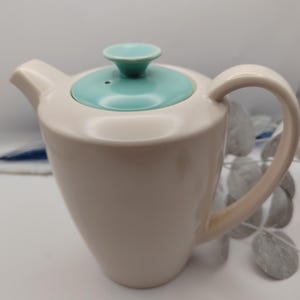 May include: Cream ceramic teapot with a turquoise lid and handle. The teapot has a classic design with a curved spout and a rounded body. The lid features a small knob. A stylish addition to any kitchen, perfect for tea.