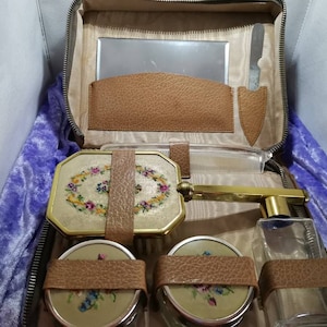 May include: A vintage brown leather travel case with a zipper closure. The case is open and contains a variety of toiletry items, including a mirror, a comb, a brush, a bottle of perfume, and a small knife. The items are all decorated with floral embroidery.