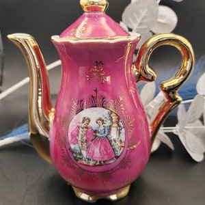 May include: A pink porcelain teapot with gold accents and a painted scene of a couple in a garden. The teapot has a lid and a spout.