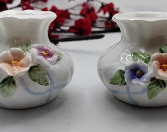May include: Two small, white ceramic vases with a bulbous shape and ruffled rims. Each vase is decorated with colourful, three-dimensional flowers in shades of purple, peach, and pink, along with green leaves. Light blue ribbons are painted on the vase.