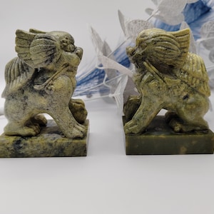 May include: A pair of green stone foo dog figurines. The figurines are sitting on square bases and are facing each other. The figurines are detailed with intricate carvings.