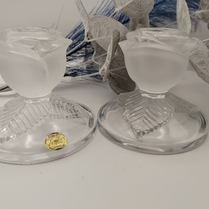 May include: A pair of clear glass candle holders shaped like roses with frosted glass petals. The holders have a leaf design on the base and a gold sticker on the bottom.