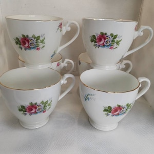 May include: Set of six white teacups with floral designs. Each cup features a pink rose with blue and white accents. The cups have a gold rim and are stacked on top of each other.