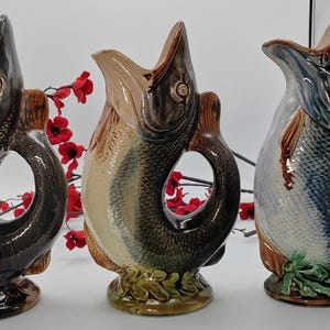 May include: Three ceramic fish-shaped jugs with different coloured glazes. The jugs are all in the same pose, with their mouths open and their tails curled around their bodies. The jugs are all about 25 cm tall.