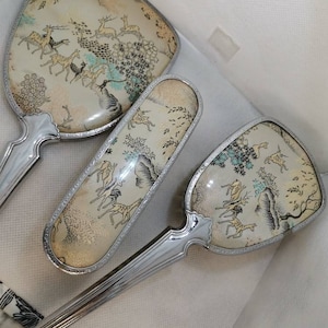 May include: A vintage silver-toned vanity set with three pieces: a hand mirror, a hairbrush, and a comb. Each piece features a delicate floral design with a light brown background and blue and green accents. The design includes stylized deer and trees.