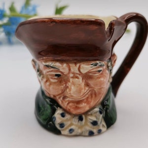 May include: A brown ceramic pitcher with a man's face and a brown hat. The pitcher has a green and white patterned design around the neck.