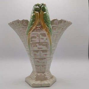 May include: A white ceramic vase with a brick pattern and two green and yellow parakeets perched on top. The vase has a wide, flared opening and a narrow base.