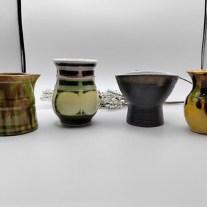 May include: Four small ceramic vases in different colors and patterns. The vases are arranged in a row on a white surface. The vase on the left is green and brown with a striped pattern. The vase in the center is green with a yellow and pink design. The vase on the right is yellow with brown streaks. The vase in the middle is black with a brown rim.