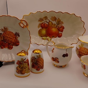 May include: A set of white porcelain serving dishes with gold trim and fruit designs. The set includes a creamer, sugar bowl, salt and pepper shakers, a small bowl, and a large serving dish.