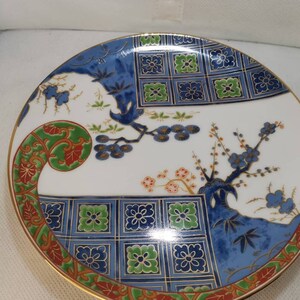 May include: A white ceramic plate with a blue, green, and gold floral design. The plate has a gold rim and is decorated with a repeating pattern of squares.