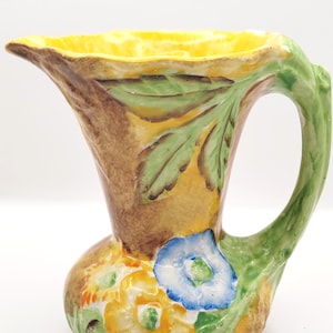 Könnte beinhalten: Ein kleiner, gelber, brauner, grüner und blauer Keramik Krug mit Blumenmuster. Der Krug hat eine breite Öffnung und einen Griff, der wie ein Ast geformt ist.