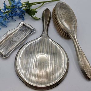 May include: A silver vanity set with a hand mirror, a brush, and a small tray. The mirror and brush have a striped pattern and the tray has a smooth surface.