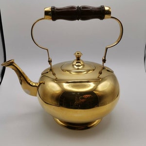 May include: A polished brass teapot with a wooden handle. The teapot has a round body and a spout on the side.