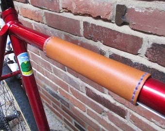 Puede incluir: Primer plano de un cuadro de bicicleta rojo con una envoltura de manillar de cuero marrón. La envoltura tiene costuras azules y está asegurada al cuadro con una abrazadera de metal. La bicicleta está apoyada contra una pared de ladrillo.