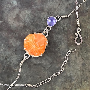 May include: A silver necklace featuring an orange seashell pendant and a purple gemstone. The necklace has a delicate chain design with a hook clasp. The seashell is the focal point, with a detailed, textured surface.