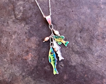 May include: A silver necklace with a pendant featuring three colorful fish charms. The fish are painted in shades of green, blue, and yellow. The necklace has a delicate chain and small pearl accents.